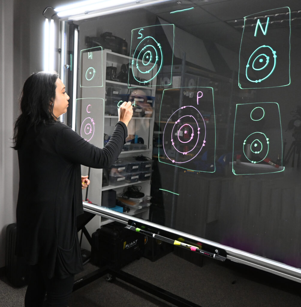 Professor writing on a light board that shows the six elements that make up life along with their Bohr models.