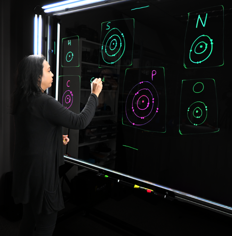 Professor writing on a light board that shows the six elements that make up life along with their Bohr models.