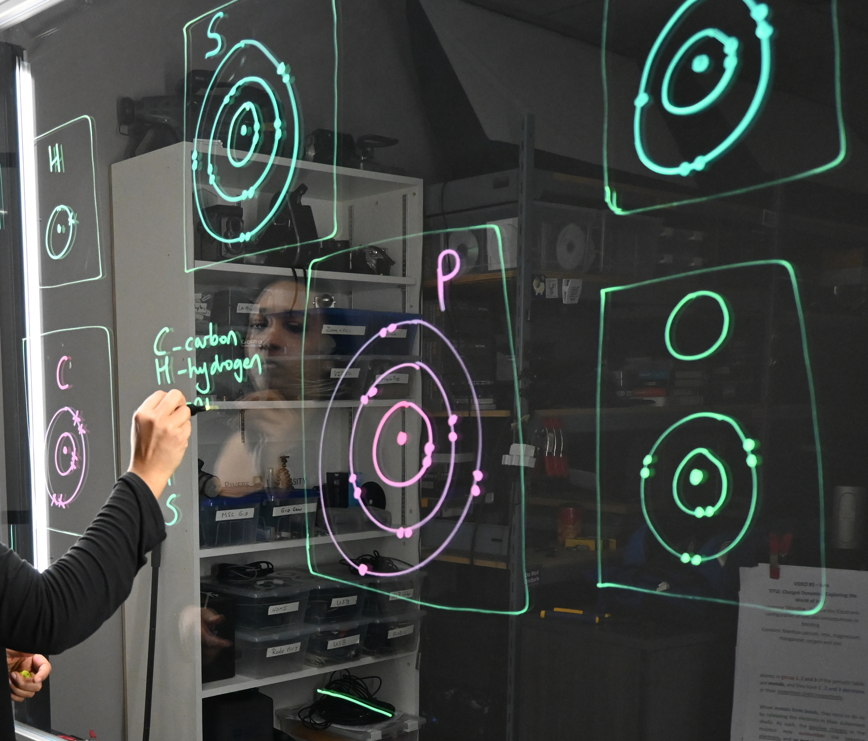 A hand writing on a light board that shows the six elements that make up life along with their Bohr models.