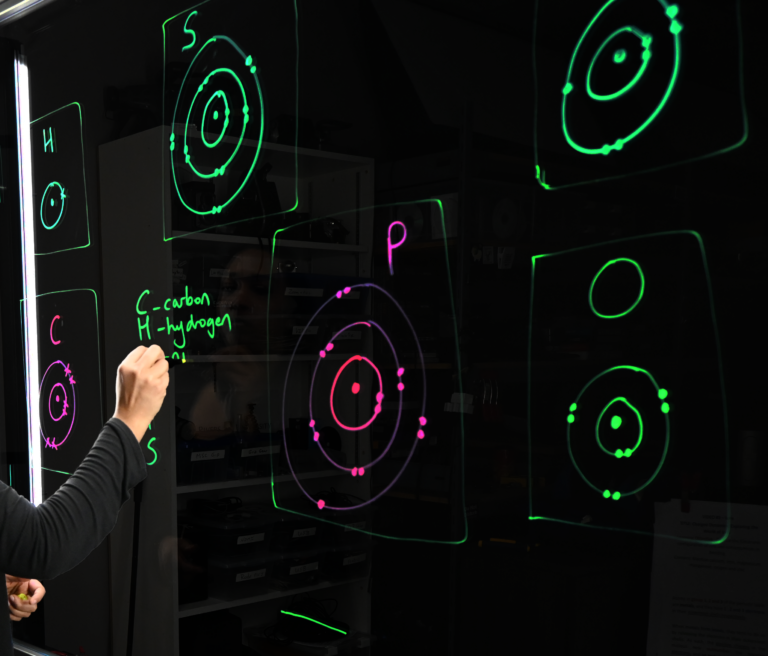 A hand writing on a light board that shows the six elements that make up life along with their Bohr models.