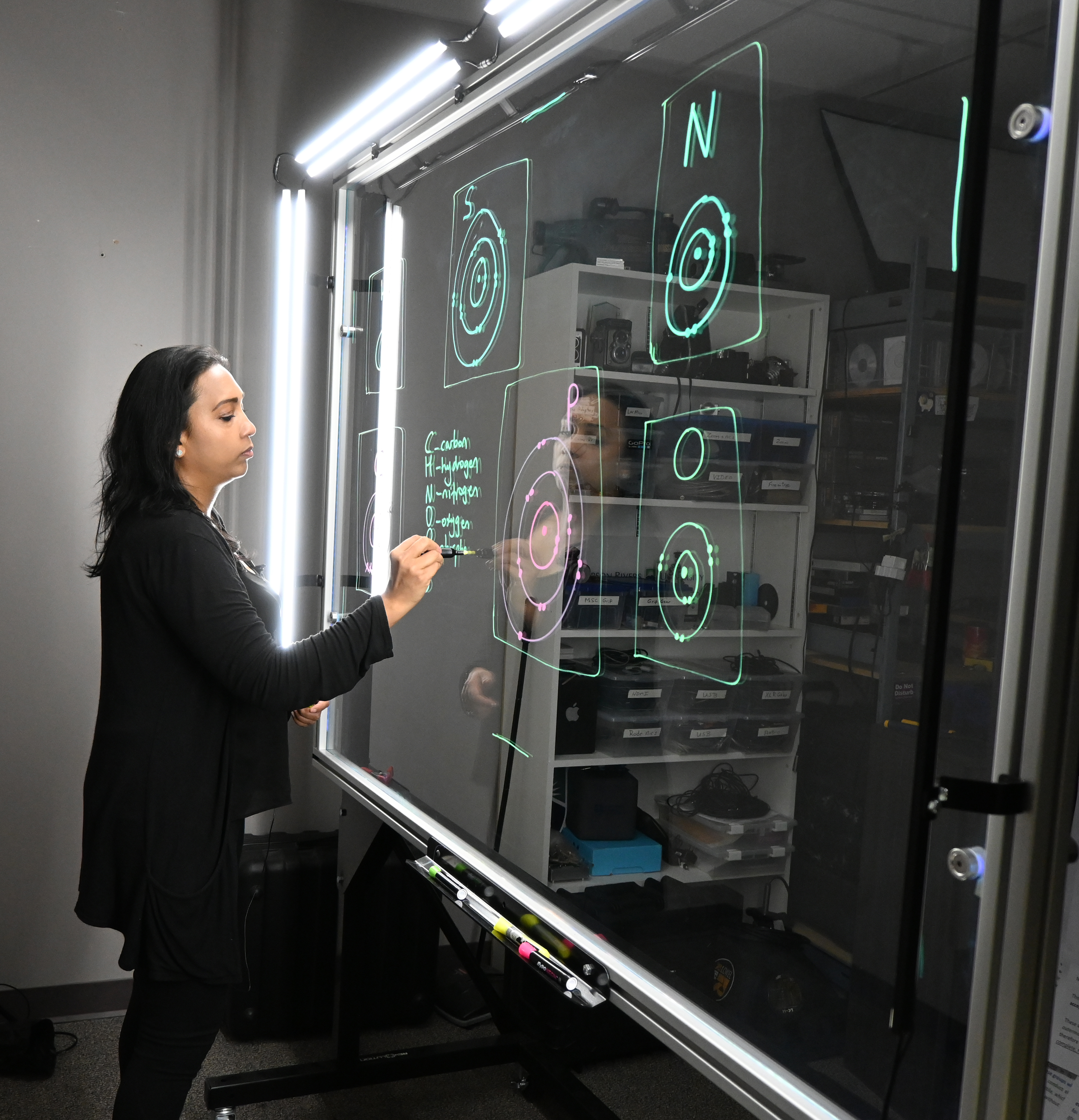 Professor writing on a light board that shows the six elements that make up life along with their Bohr models.