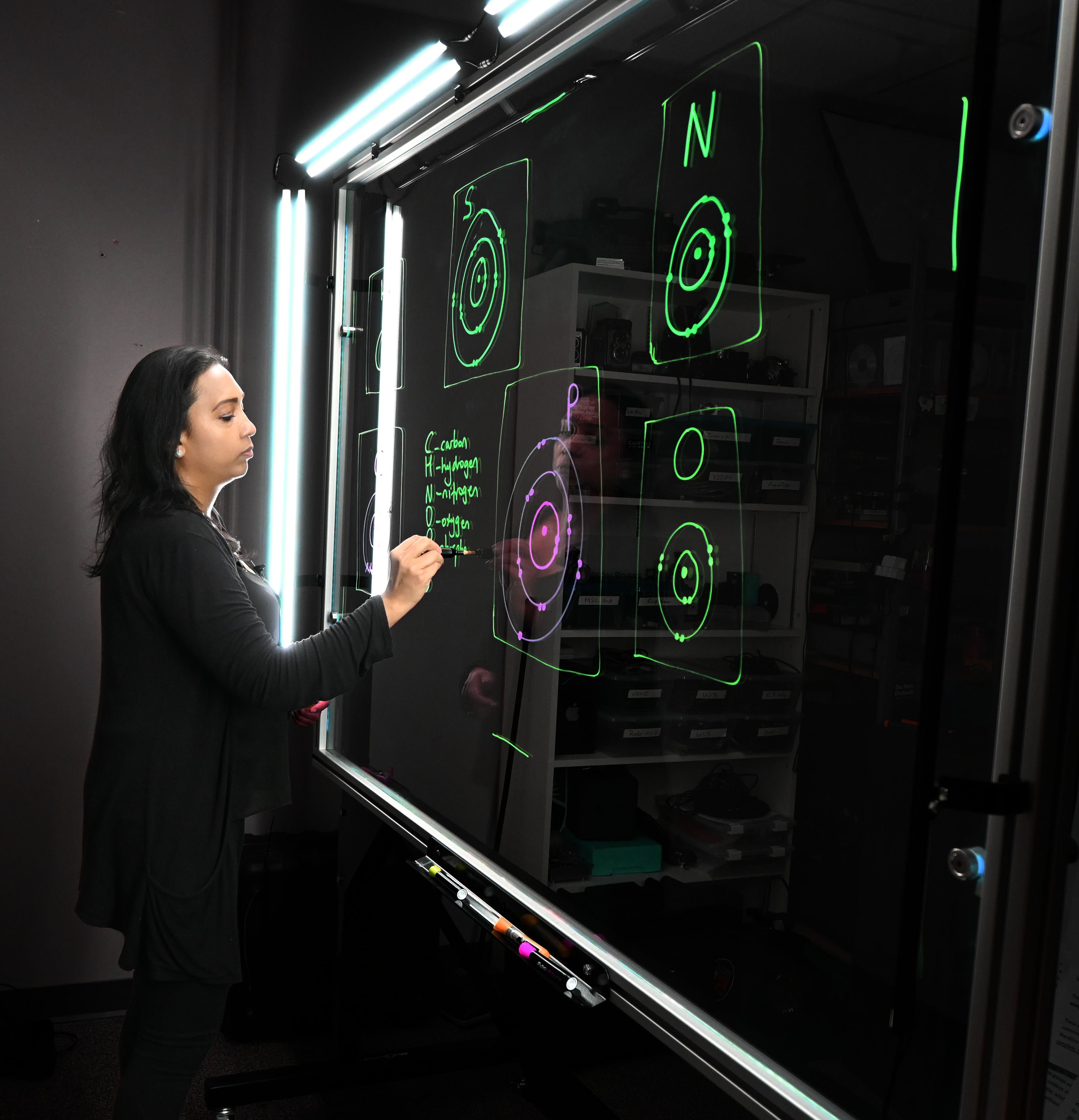 Professor writing on a light board that shows the six elements that make up life along with their Bohr models.