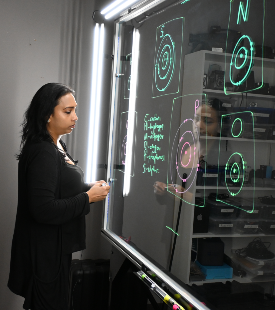 Professor reading writing on a light board that shows the six elements that make up life along with their Bohr models.