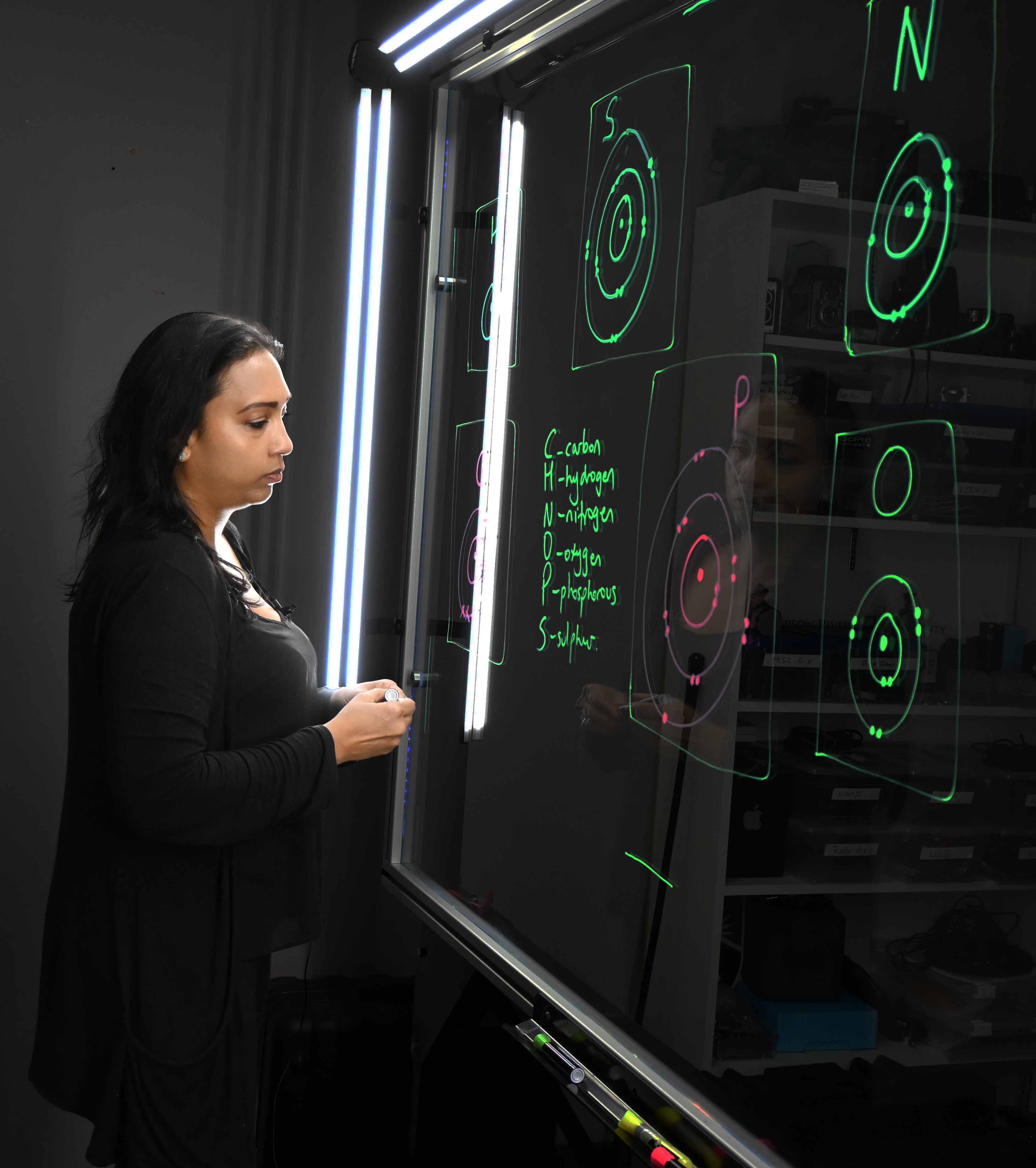 Professor reading writing on a light board that shows the six elements that make up life along with their Bohr models.