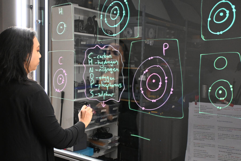 Professor writing on a light board that shows the six elements that make up life along with their Bohr models.