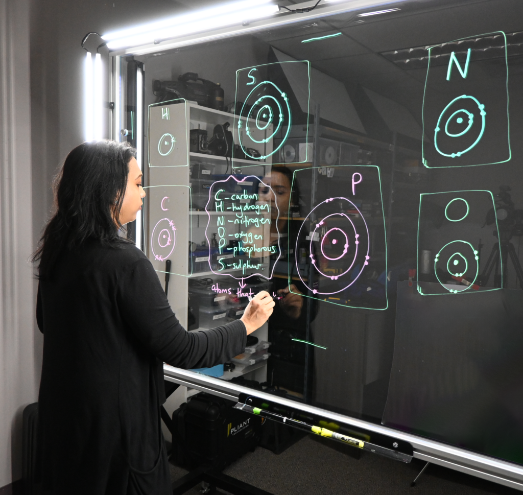 Professor writing on a light board that shows the six elements that make up life along with their Bohr models.