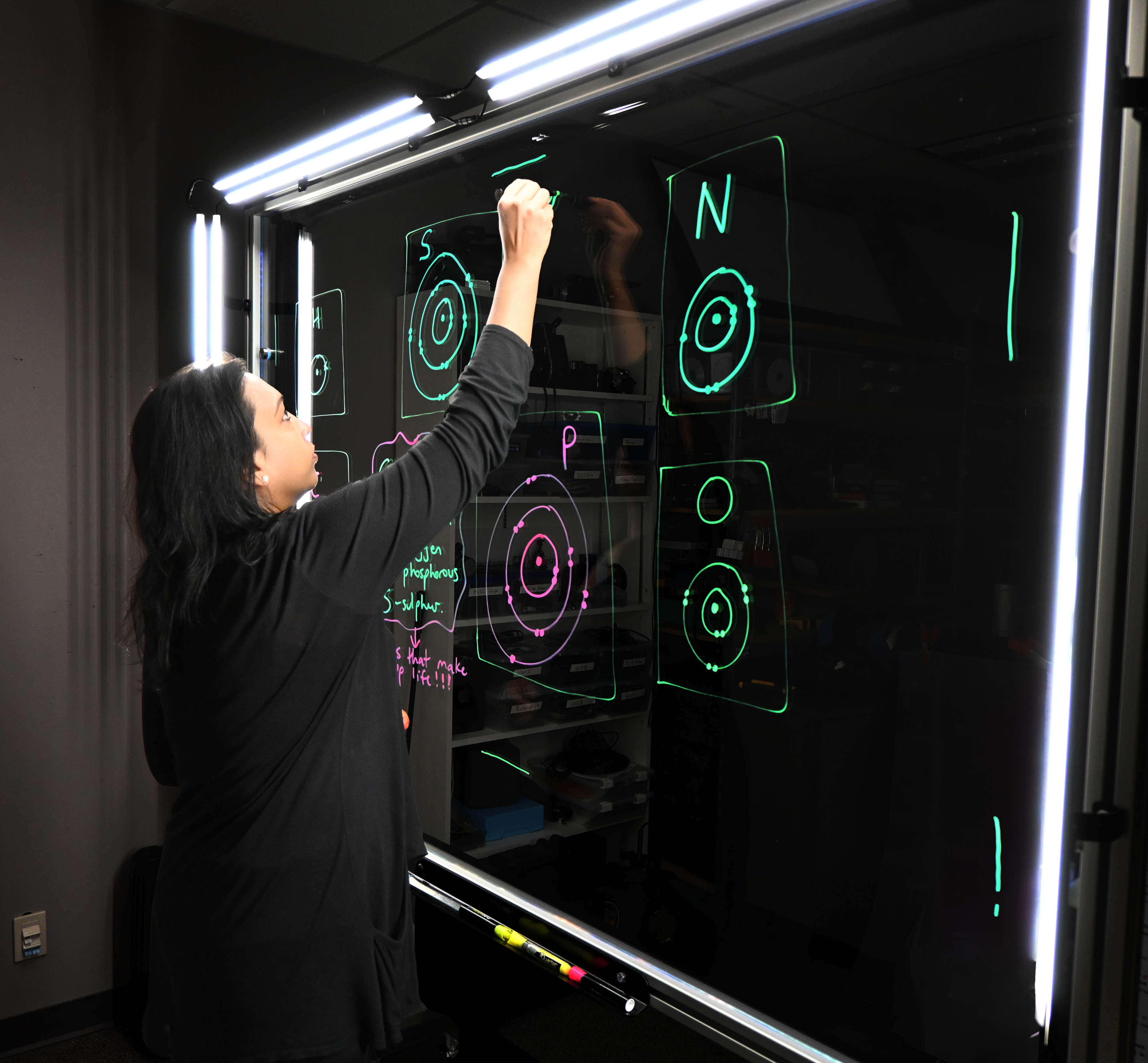 Professor writing on a light board that shows the six elements that make up life along with their Bohr models.