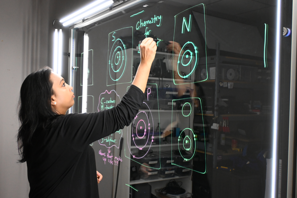 Professor writing on a light board that shows the six elements that make up life along with their Bohr models.