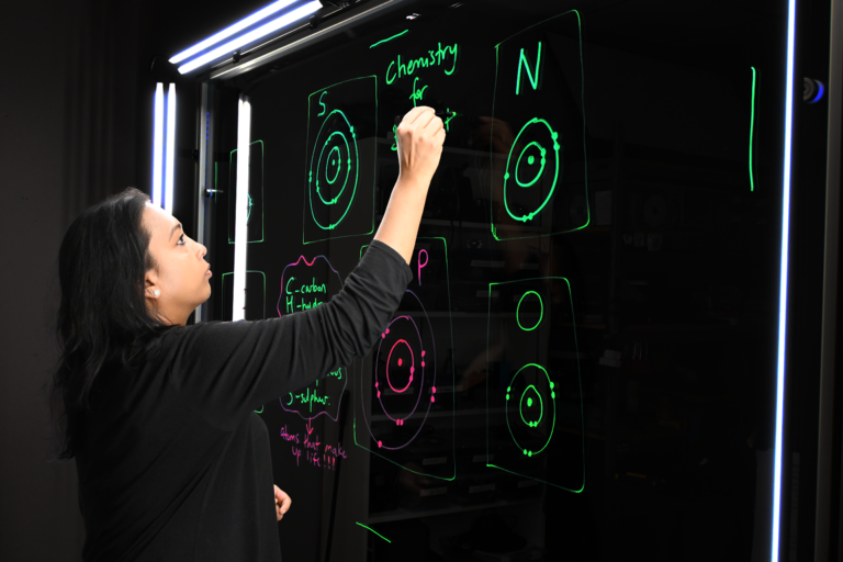 Professor writing on a light board that shows the six elements that make up life along with their Bohr models.