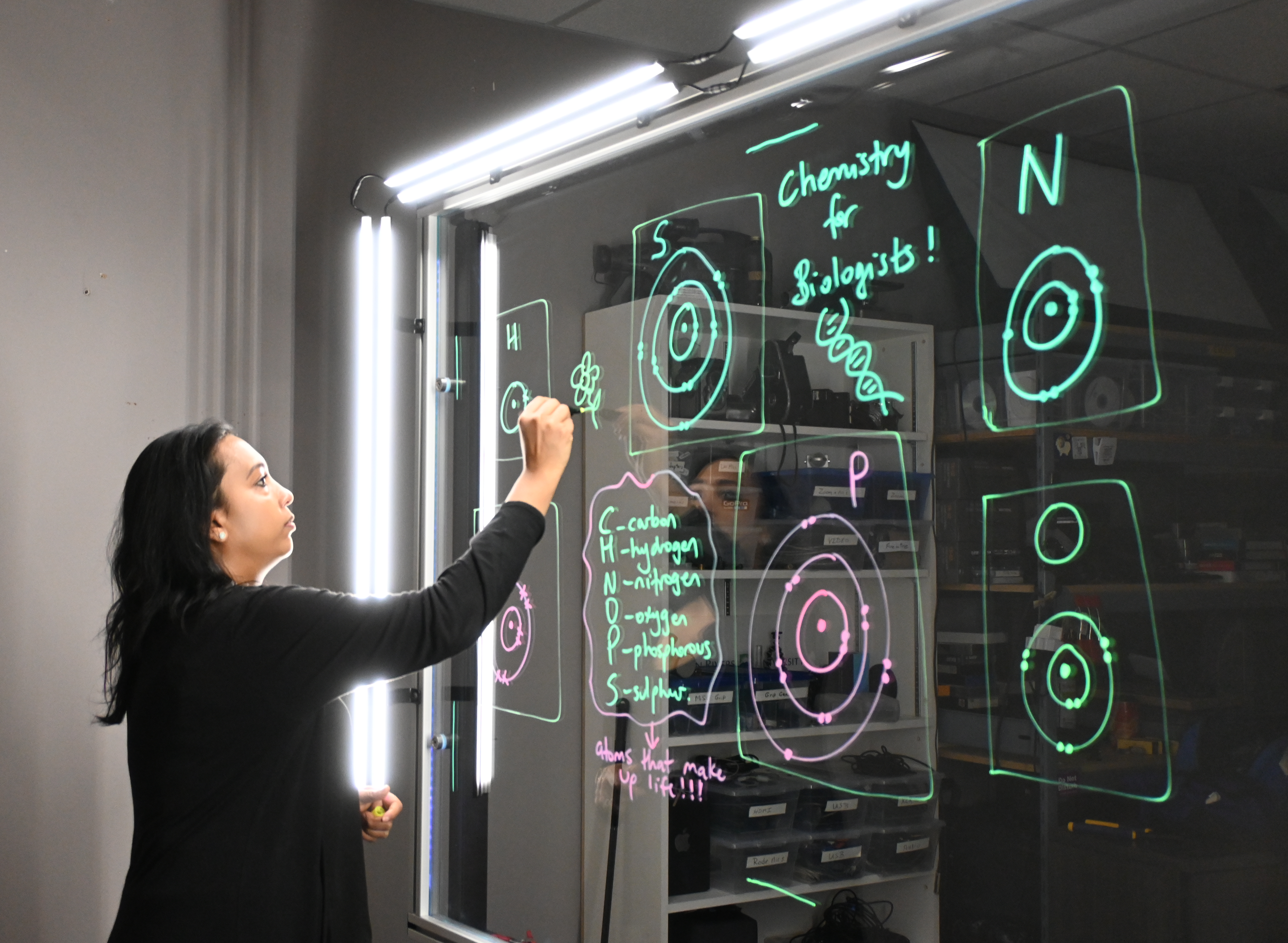 Professor writing on a light board that shows the six elements that make up life along with their Bohr models.