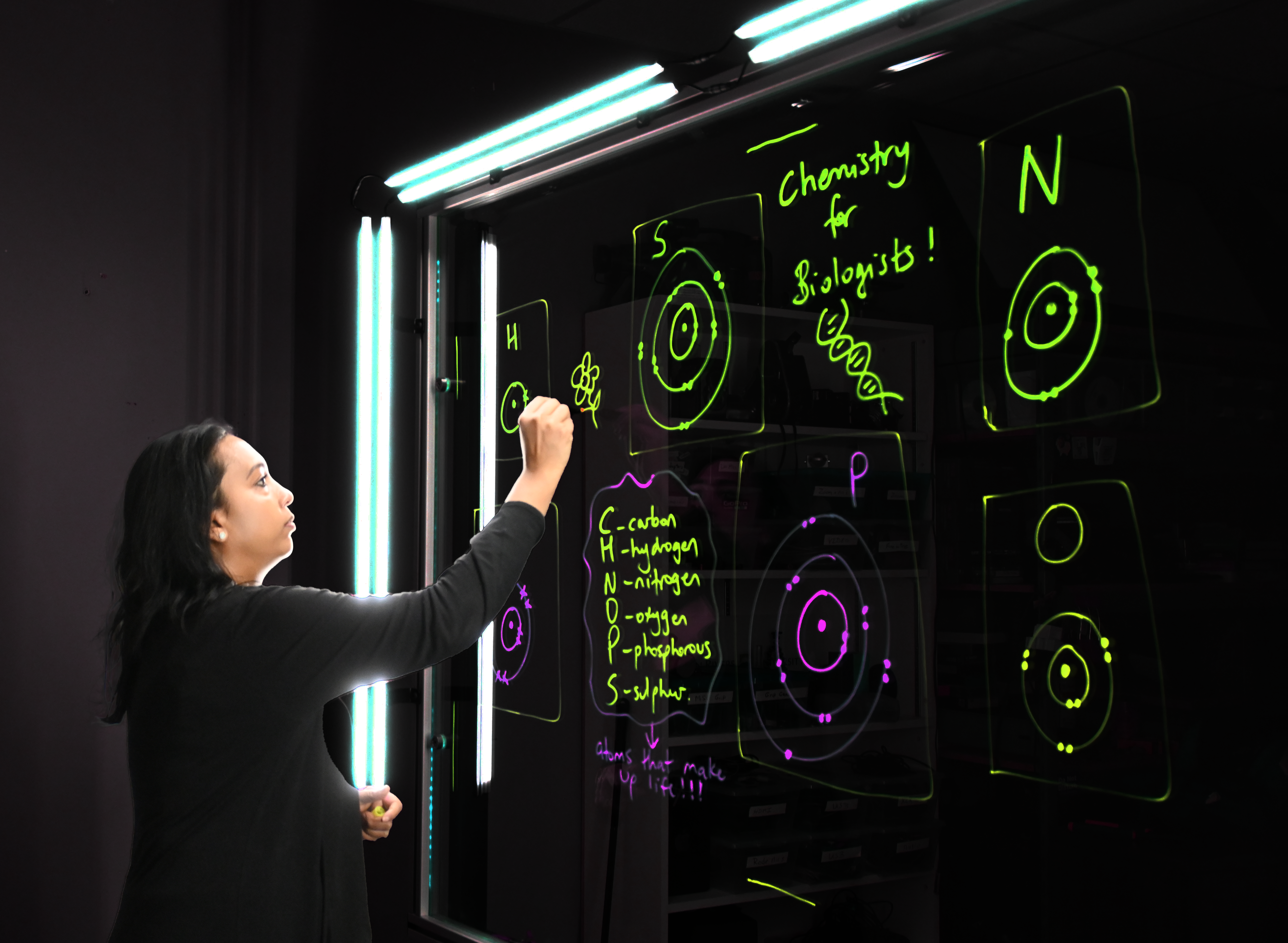 Professor writing on a light board that shows the six elements that make up life along with their Bohr models.