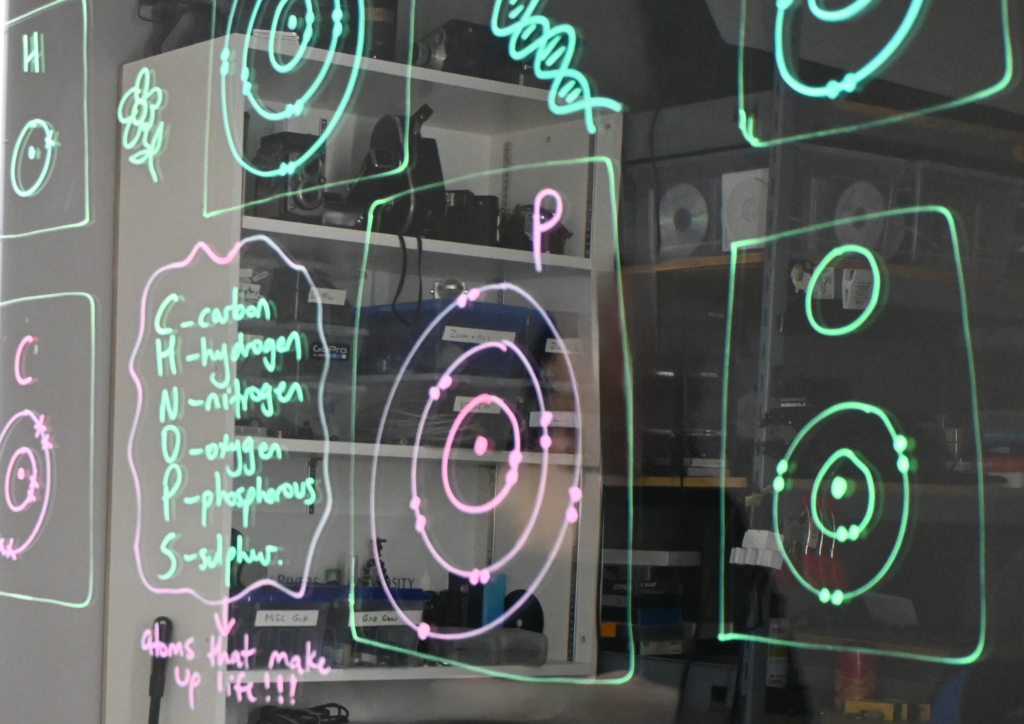 Writing on a light board shows Bohr models of sulphur, nitrogen, phosphorus, and oxygen. There is a list of the six elements that make up life: carbon, hydrogen, nitrogen, oxygen, phosphorus, and sulphur.