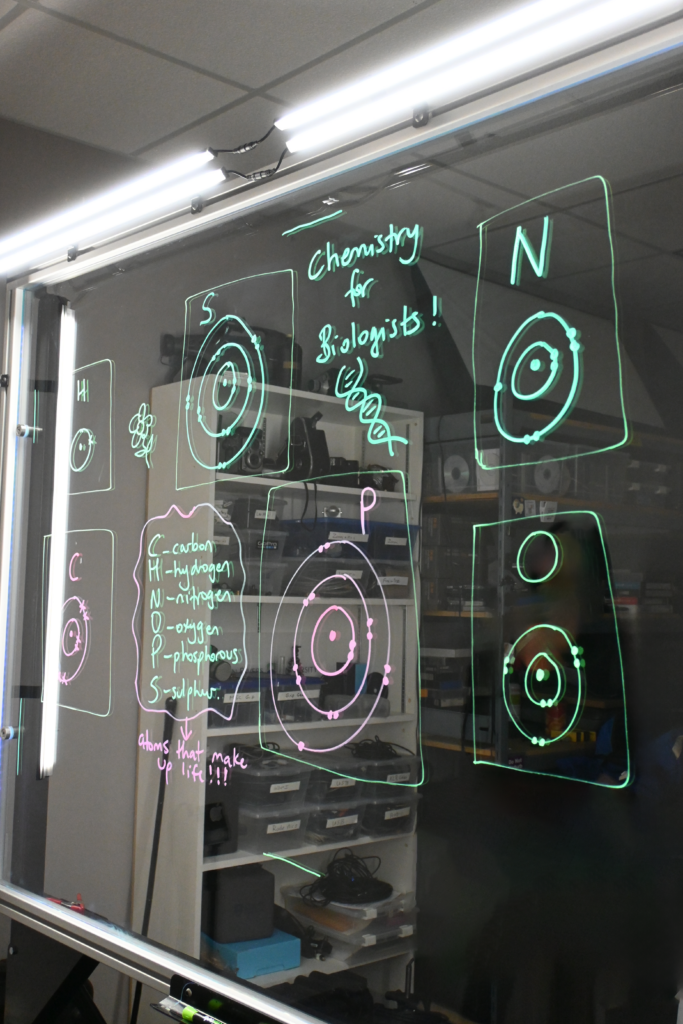 Writing on a light board says 'Chemistry for Biologists' and lists the six elements that make up life along with their Bohr models.