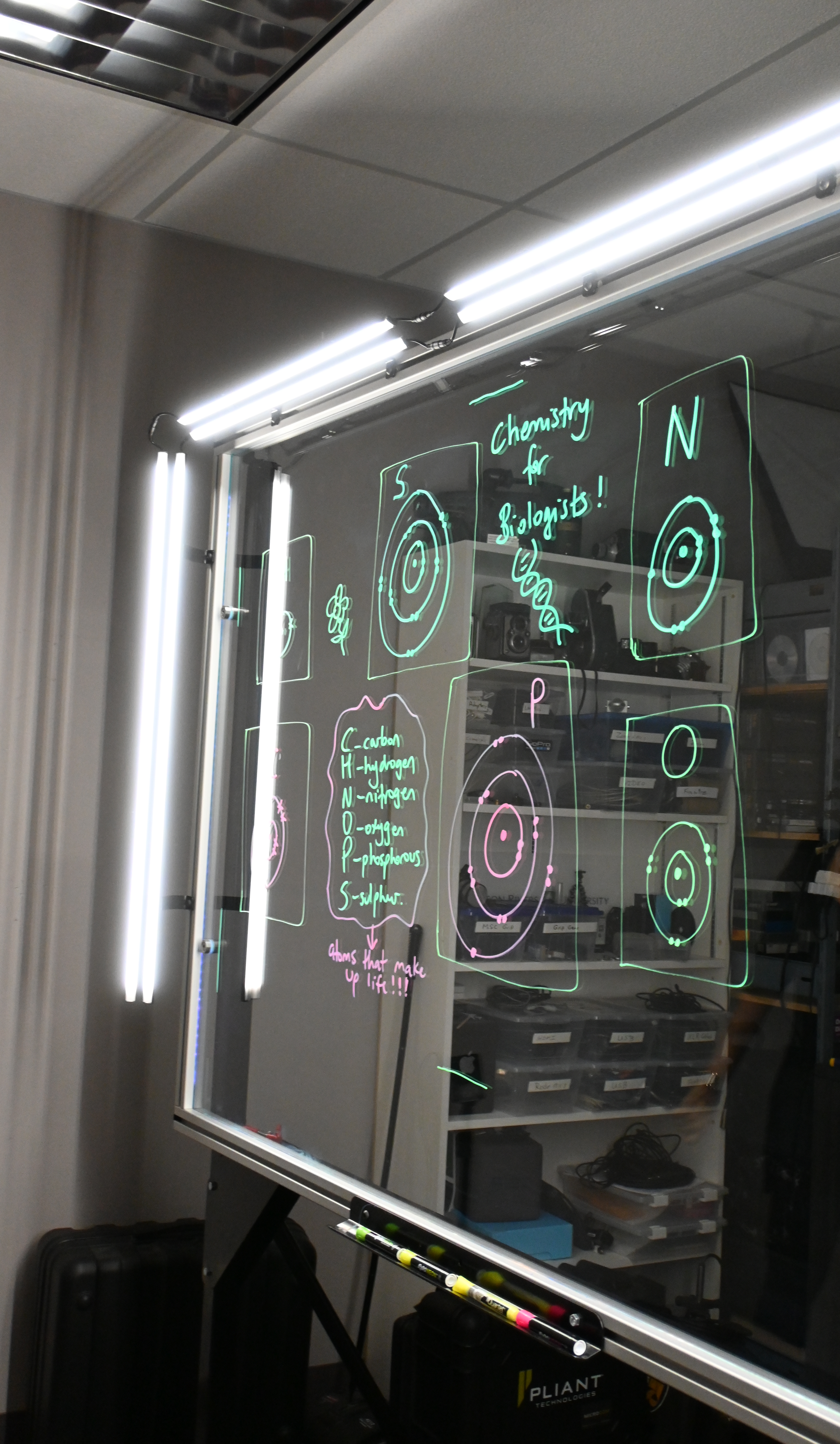 Writing on a light board says 'Chemistry for Biologists' and lists the six elements that make up life along with their Bohr models.