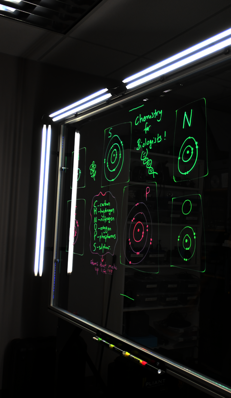 Writing on a light board says 'Chemistry for Biologists' and lists the six elements that make up life along with their Bohr models.