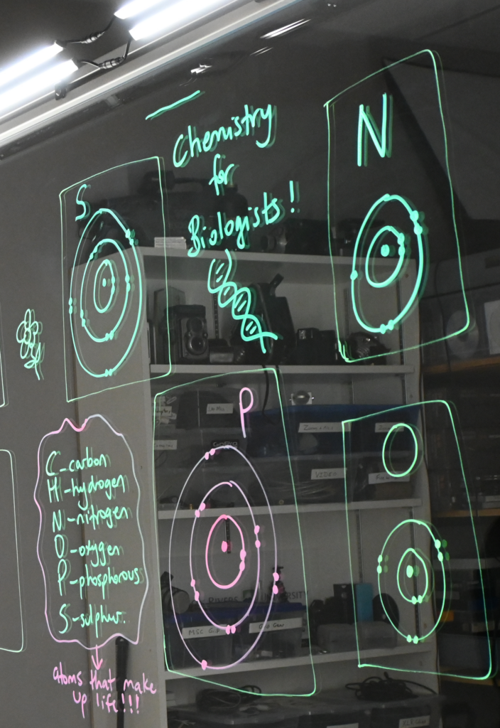 Writing on a light board shows Bohr models of sulphur, nitrogen, phosphorus, and oxygen. There is a list of the six elements that make up life: carbon, hydrogen, nitrogen, oxygen, phosphorus, and sulphur.