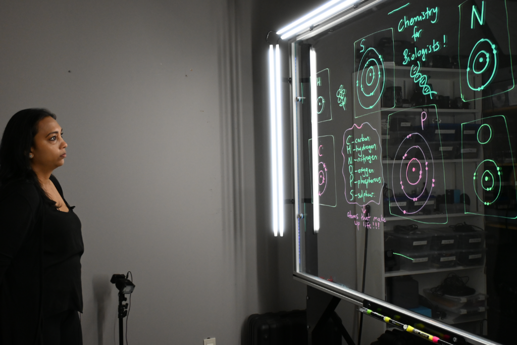 Professor looking at a light board that shows a list of the six elements that make up life along with their Bohr models.