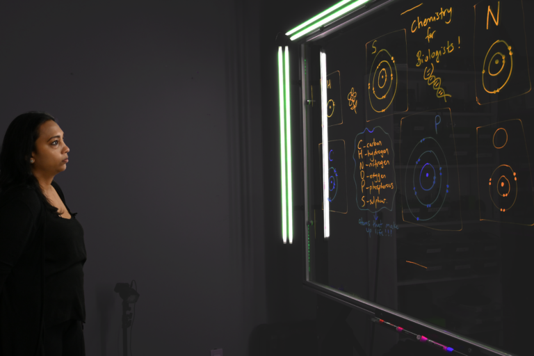 Professor looking at a light board that shows a list of the six elements that make up life along with their Bohr models.