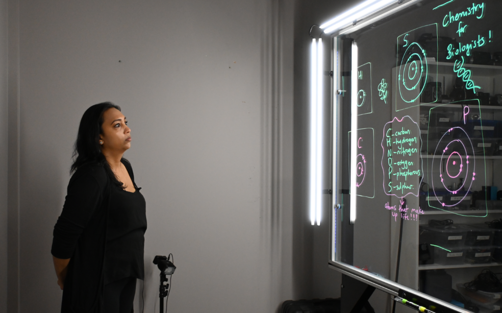 Professor looking at a light board that shows a list of the six elements that make up life along with their Bohr models.