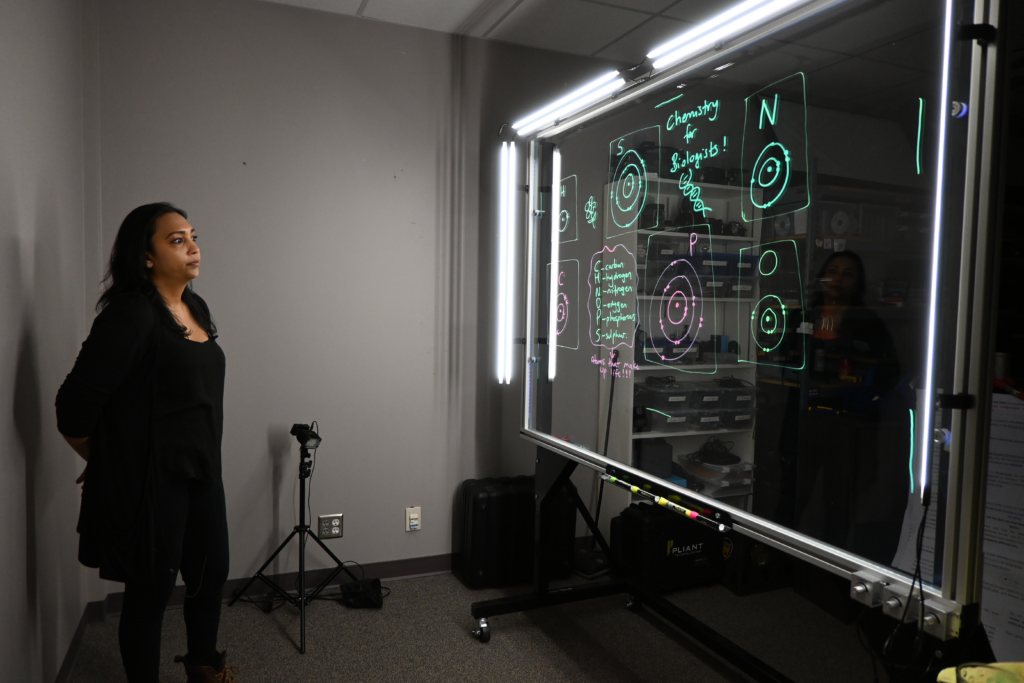 Professor looking at a light board that shows a list of the six elements that make up life along with their Bohr models.