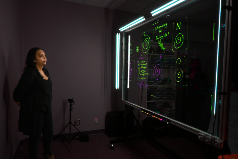 Professor looking at a light board that shows a list of the six elements that make up life along with their Bohr models.