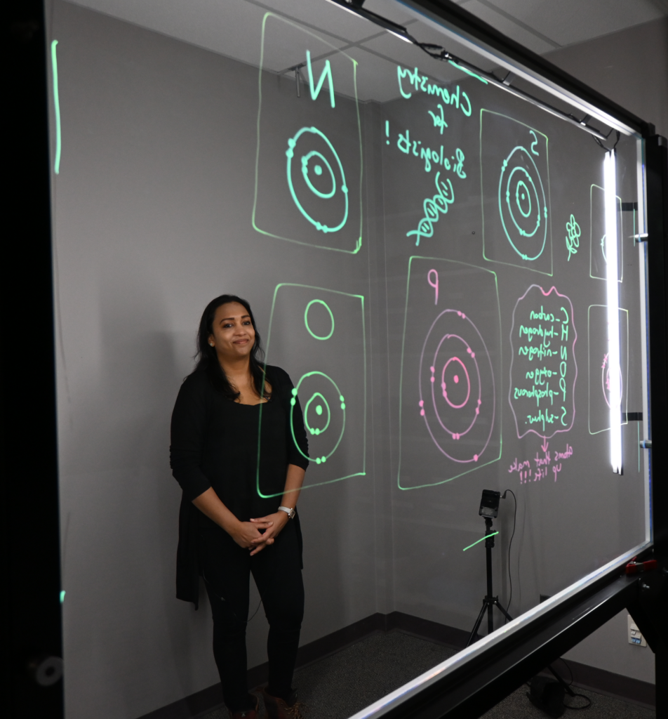 Professor standing behind a light board that shows a list of the six elements that make up life along with their Bohr models.