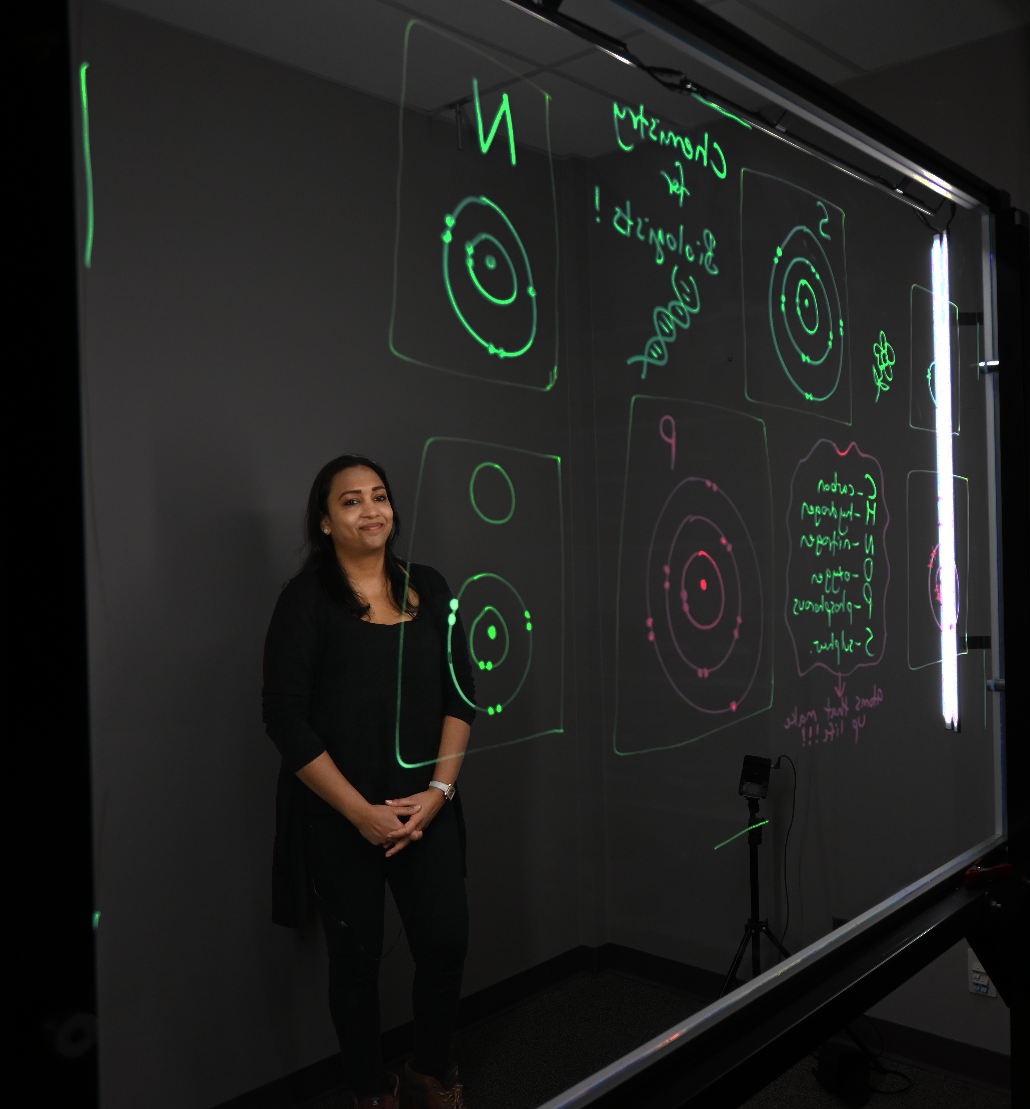 Professor standing behind a light board that shows a list of the six elements that make up life along with their Bohr models.