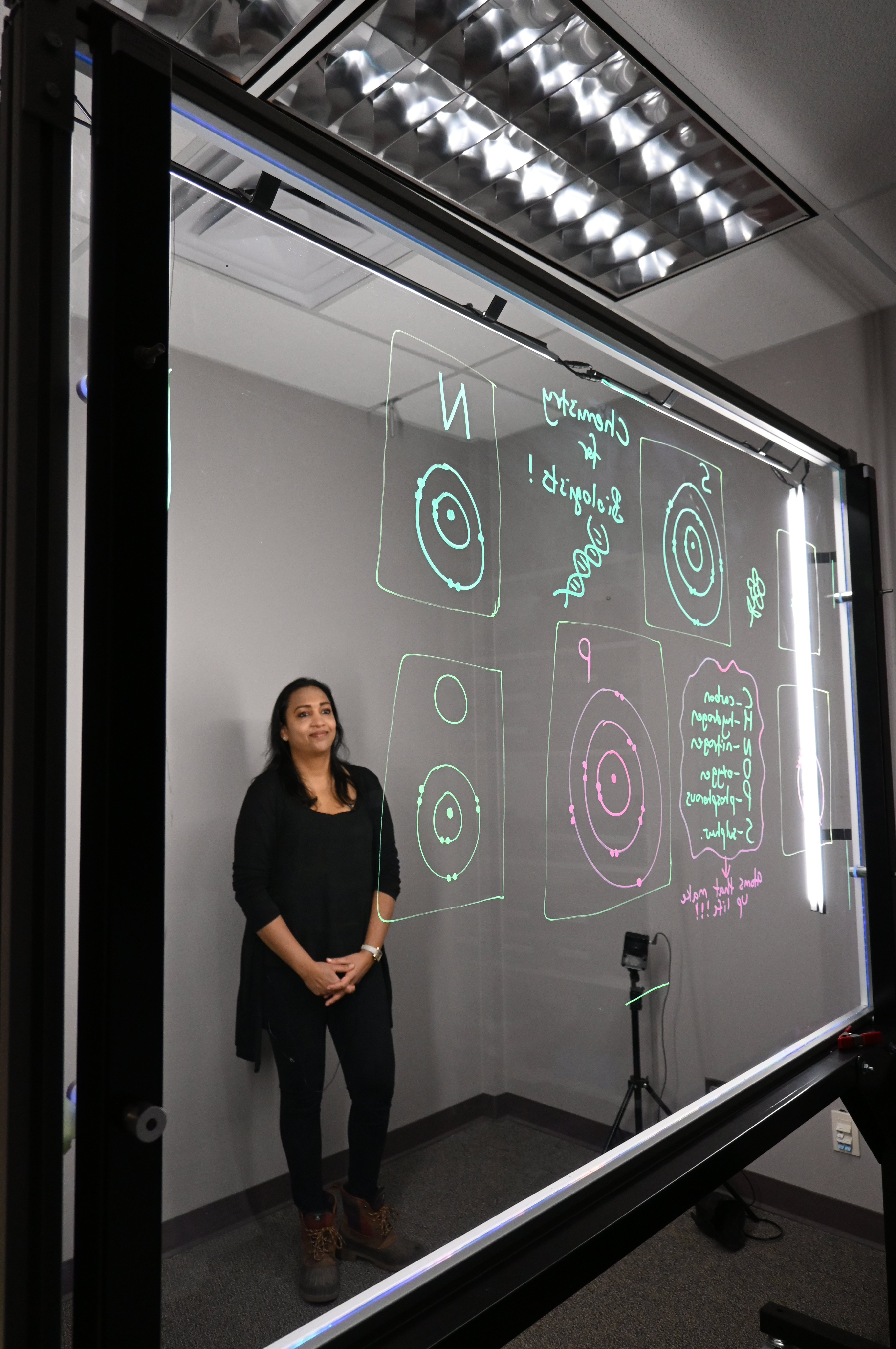 Professor standing behind a light board that shows a list of the six elements that make up life along with their Bohr models.