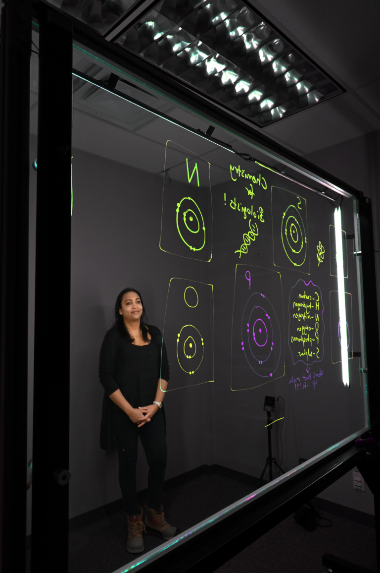 Professor standing behind a light board that shows a list of the six elements that make up life along with their Bohr models.