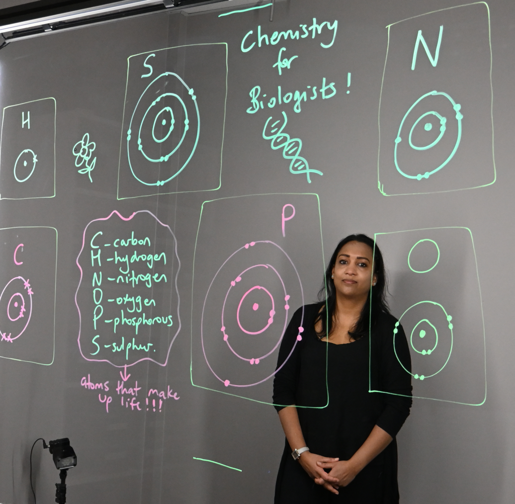 Professor standing behind a light board that shows a list of the six elements that make up life along with their Bohr models.