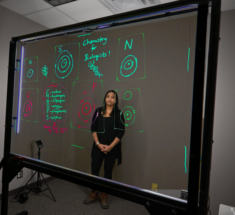 Professor standing behind a light board that shows a list of the six elements that make up life along with their Bohr models.