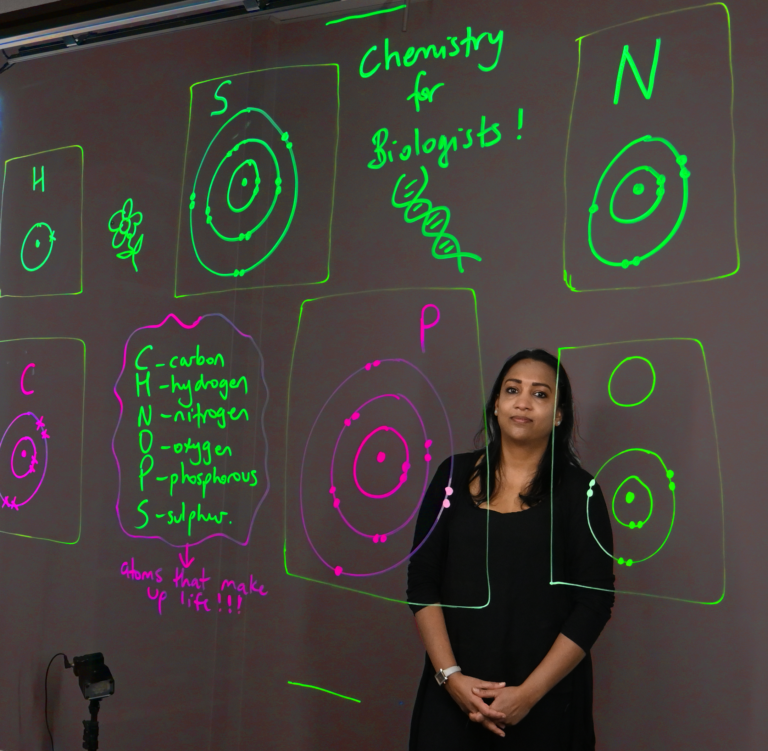 Professor standing behind a light board that shows a list of the six elements that make up life along with their Bohr models.