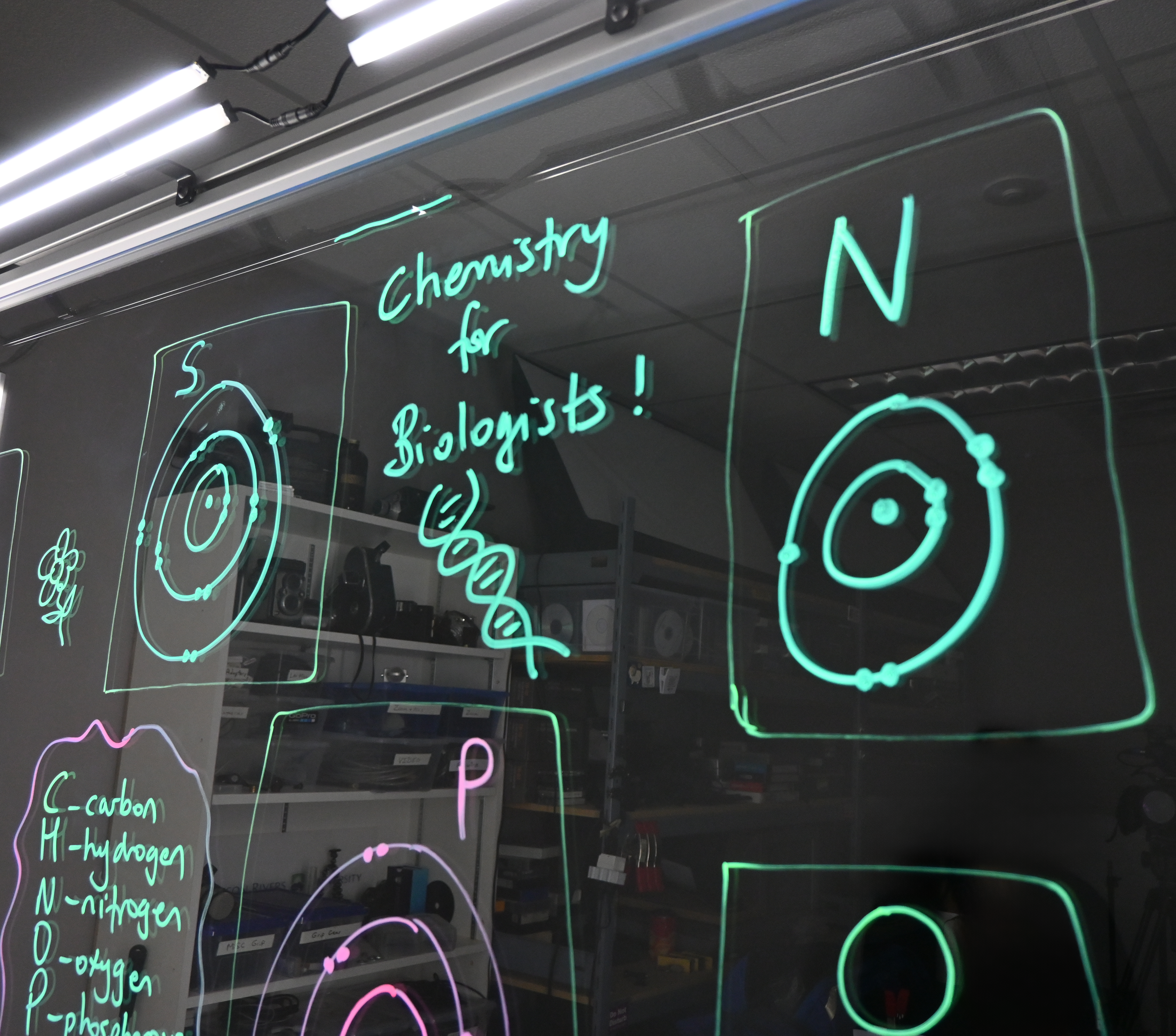 Writing on a light board says 'Chemistry for Biologists' with a double helix below it. Around the writing are Bohr models of sulphur, nitrogen, and phosphorus.