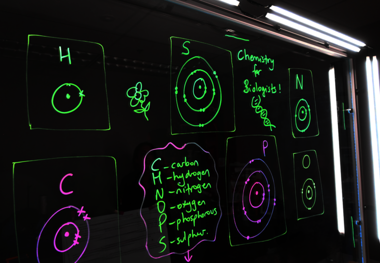 Writing on a light board says 'Chemistry for Biologists' and lists the six elements that make up life along with their Bohr models.