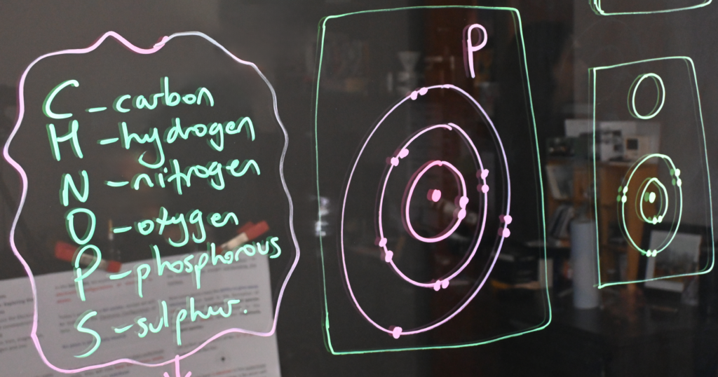 Writing on a light board shows Bohr models of phosphorus and oxygen and lists the six elements that make up life. These elements are carbon, hydrogen, nitrogen, oxygen, phosphorus, and sulphur.