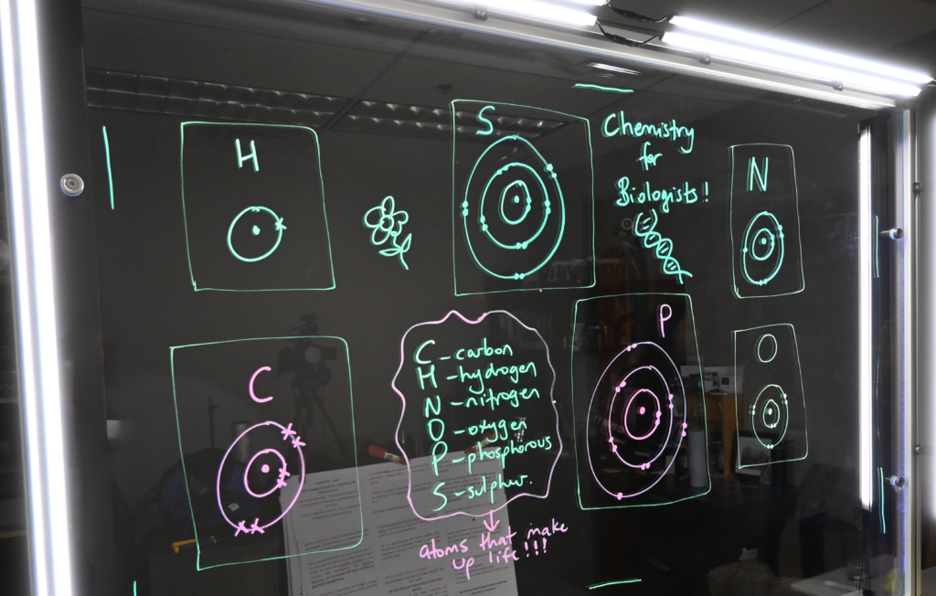 Writing on a light board says 'Chemistry for Biologists' and lists the six elements that make up life along with their Bohr models.