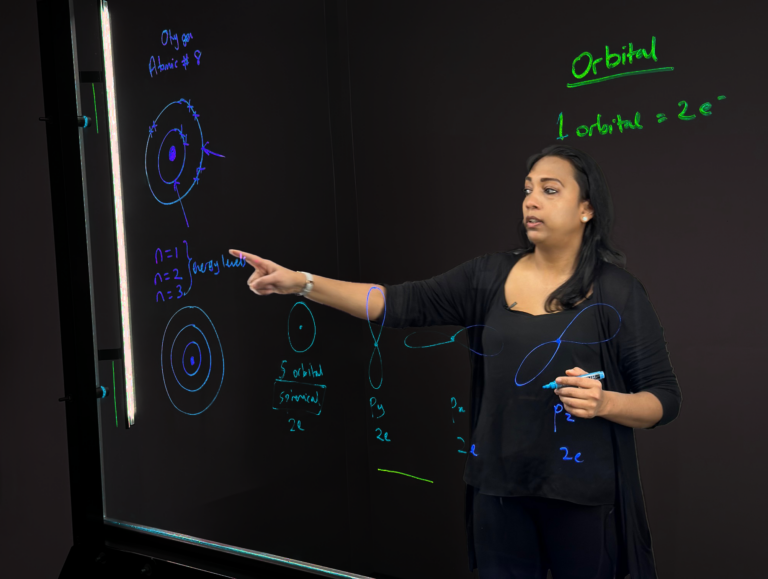 Professor pointing to to writing on a light board that shows a Bohr model of the element oxygen.